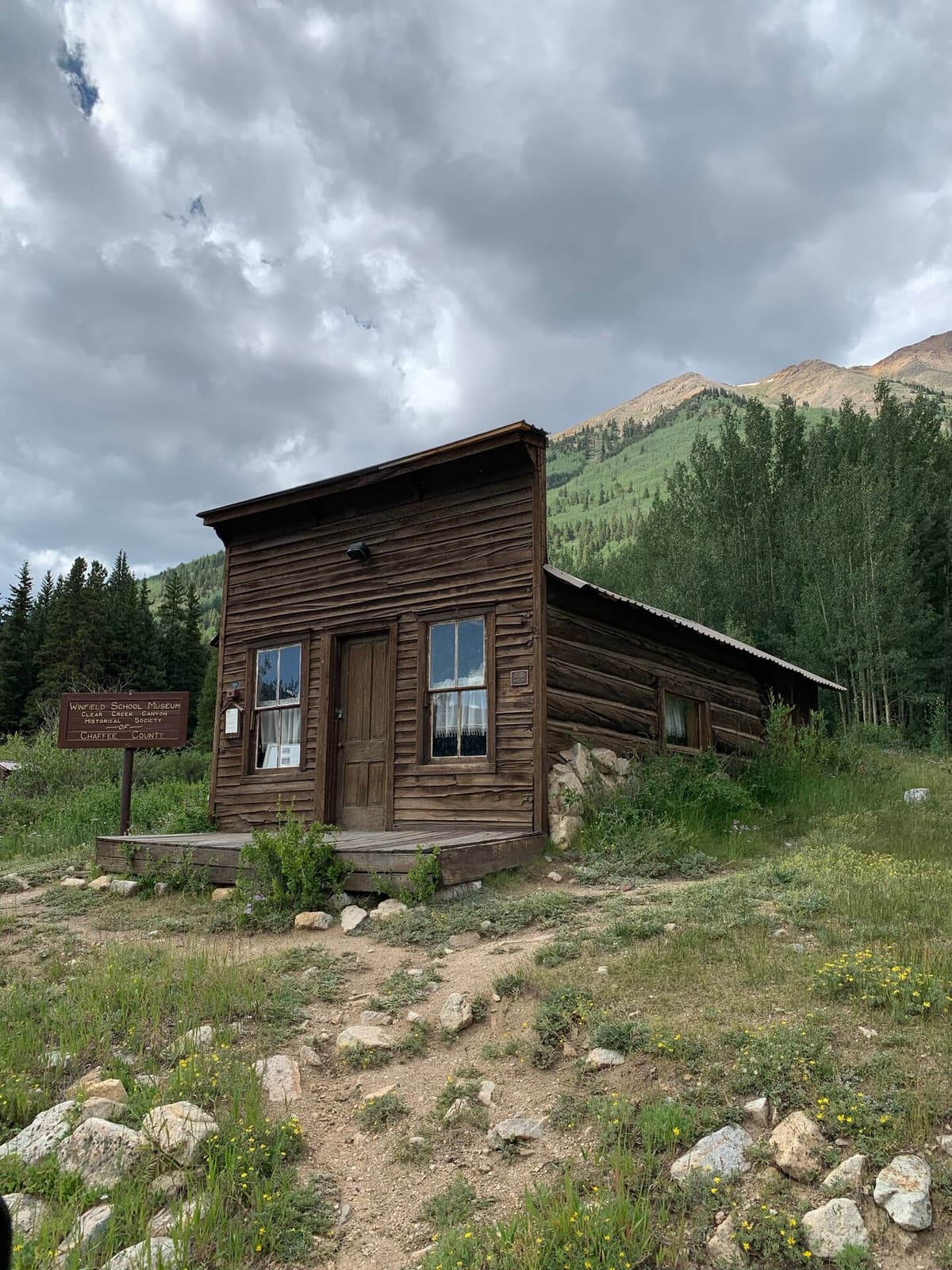 The restored wooden false-front Winfield schoolhouse museum standing in a green mountain meadow with peaks behind, in the Colorado ghost town of Winfield